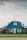 A blue house with a triangular roof and white-framed windows is centered in the image. The house has a small, fenced front yard with a well-maintained lawn. There are a few leafless trees and shrubs near the entrance. The overcast sky provides a muted backdrop to the setting.