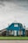 A blue house with a triangular roof and white-framed windows is centered in the image. The house has a small, fenced front yard with a well-maintained lawn. There are a few leafless trees and shrubs near the entrance. The overcast sky provides a muted backdrop to the setting.