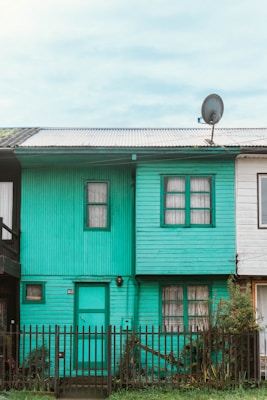 A small teal-colored wooden house with two floors featuring a corrugated metal roof and a satellite dish attached to it. There are two windows on the upper floor and a door with a window beside it on the ground floor. The house is flanked by two other houses on either side and a small black picket fence in front.