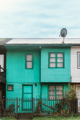 A small teal-colored wooden house with two floors featuring a corrugated metal roof and a satellite dish attached to it. There are two windows on the upper floor and a door with a window beside it on the ground floor. The house is flanked by two other houses on either side and a small black picket fence in front.