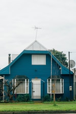 A blue house with a steeply pitched roof features large front-facing windows and a central door. There is a fenced front yard with a well-maintained lawn and some plants near the entrance. An antenna is mounted at the top of the roof, and there is a satellite dish on the side.