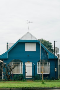 A blue house with a steeply pitched roof features large front-facing windows and a central door. There is a fenced front yard with a well-maintained lawn and some plants near the entrance. An antenna is mounted at the top of the roof, and there is a satellite dish on the side.
