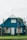 A blue house with a steeply pitched roof features large front-facing windows and a central door. There is a fenced front yard with a well-maintained lawn and some plants near the entrance. An antenna is mounted at the top of the roof, and there is a satellite dish on the side.