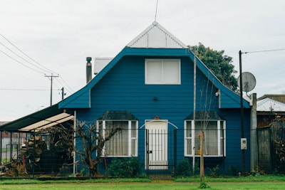 A quaint blue house with a steep roof and a central white door is surrounded by a wooden fence. The house features two windows flanking the entrance and a chimney peeking at the back. An attached open shelter on the side houses logs and various items.