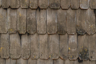 A close-up view of a rustic wooden shingle roof with weathered, overlapping wooden tiles. The surface texture displays signs of aging and wear, with some lichen or moss growth in patches.
