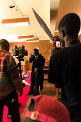 A film set scene with a group of people, including one person who appears to be acting, surrounded by cameras and lighting equipment. The setting has a warm, ambient lighting with decorations in the background and a red carpet on the floor.