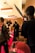 A film set scene with a group of people, including one person who appears to be acting, surrounded by cameras and lighting equipment. The setting has a warm, ambient lighting with decorations in the background and a red carpet on the floor.