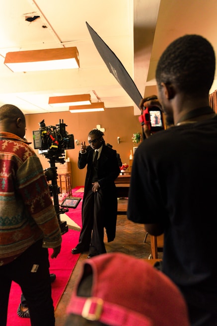 A film set scene with a group of people, including one person who appears to be acting, surrounded by cameras and lighting equipment. The setting has a warm, ambient lighting with decorations in the background and a red carpet on the floor.