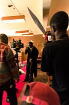 A film set scene with a group of people, including one person who appears to be acting, surrounded by cameras and lighting equipment. The setting has a warm, ambient lighting with decorations in the background and a red carpet on the floor.