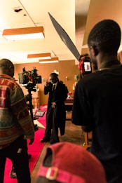 A film set scene with a group of people, including one person who appears to be acting, surrounded by cameras and lighting equipment. The setting has a warm, ambient lighting with decorations in the background and a red carpet on the floor.