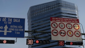 Several traffic signs are displayed against the backdrop of a modern glass building. A blue sign indicates directions to Sala with distances, while a red and white sign prohibits certain activities such as walking and features symbols for road restrictions. There are digital displays showing the number 38.