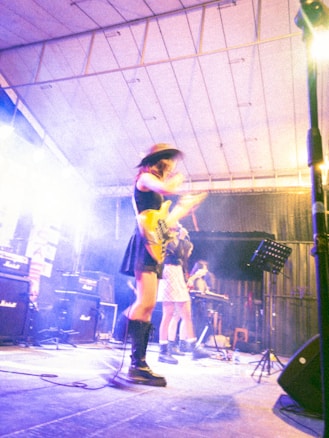 A band performs on a stage with vibrant lighting. The musicians appear absorbed in their music, with one playing a guitar and another possibly on keyboard. They wear stylish outfits, including hats and boots. Amplifiers are visible in the background, adding to the concert atmosphere.
