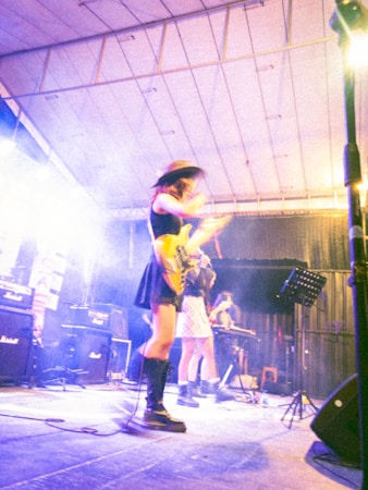 A band performs on a stage with vibrant lighting. The musicians appear absorbed in their music, with one playing a guitar and another possibly on keyboard. They wear stylish outfits, including hats and boots. Amplifiers are visible in the background, adding to the concert atmosphere.