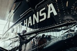 A close-up view of a metallic vehicle surface with rivets. The word 'HANSA' is prominently displayed in large white letters. The reflection on the surface suggests it could be an aircraft or a similar type of vehicle.
