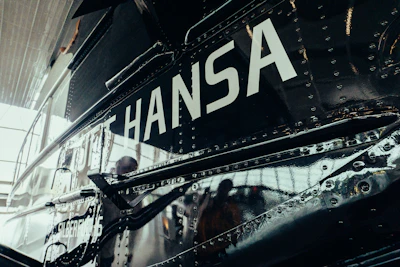 A close-up view of a metallic vehicle surface with rivets. The word 'HANSA' is prominently displayed in large white letters. The reflection on the surface suggests it could be an aircraft or a similar type of vehicle.