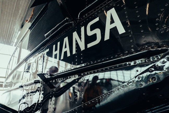 A close-up view of a metallic vehicle surface with rivets. The word 'HANSA' is prominently displayed in large white letters. The reflection on the surface suggests it could be an aircraft or a similar type of vehicle.