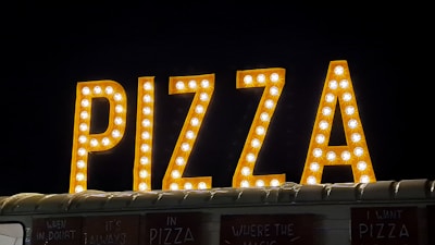 Large, illuminated letters spelling 'PIZZA' are prominently displayed against a dark background. Each letter is filled with round light bulbs, giving off a bright and inviting appearance. Below the sign, a part of a food truck or stall is visible.
