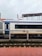 A train carriage is stationed on the tracks at a railway platform. The carriage is primarily white with a blue stripe and features the Indian Railways logo. There is a small Indian flag above one of the windows. The platform has red tiles, and there are overhead lines visible, indicating electric tracks.