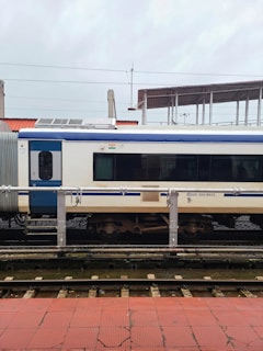 A train carriage is stationed on the tracks at a railway platform. The carriage is primarily white with a blue stripe and features the Indian Railways logo. There is a small Indian flag above one of the windows. The platform has red tiles, and there are overhead lines visible, indicating electric tracks.