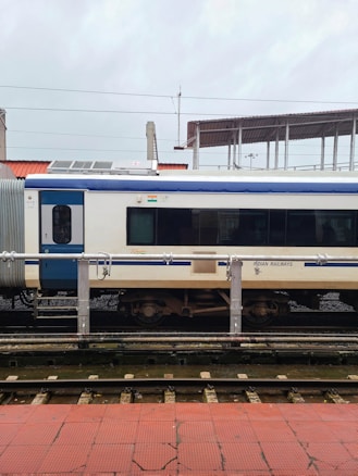A train carriage is stationed on the tracks at a railway platform. The carriage is primarily white with a blue stripe and features the Indian Railways logo. There is a small Indian flag above one of the windows. The platform has red tiles, and there are overhead lines visible, indicating electric tracks.