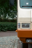 An up-close view of the front part of a parked vehicle, showcasing its headlight and bumper. The vehicle is two-toned, with a beige upper section and an orange lower section. A sticker with cartoon ears is visible on the windshield. The vehicle is parked on cobblestone pavement with lush greenery in the background.