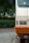 An up-close view of the front part of a parked vehicle, showcasing its headlight and bumper. The vehicle is two-toned, with a beige upper section and an orange lower section. A sticker with cartoon ears is visible on the windshield. The vehicle is parked on cobblestone pavement with lush greenery in the background.