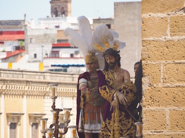 A detailed procession display featuring religious statues dressed in ornate garments. The central figure wears a purple robe with gold embroidery, crowned with a headpiece of feathers, symbolizing a sacred or historical event. A Roman soldier figure is adorned in armor with a striking feathered helmet. The background reveals a cityscape with a mix of modern and traditional architecture.