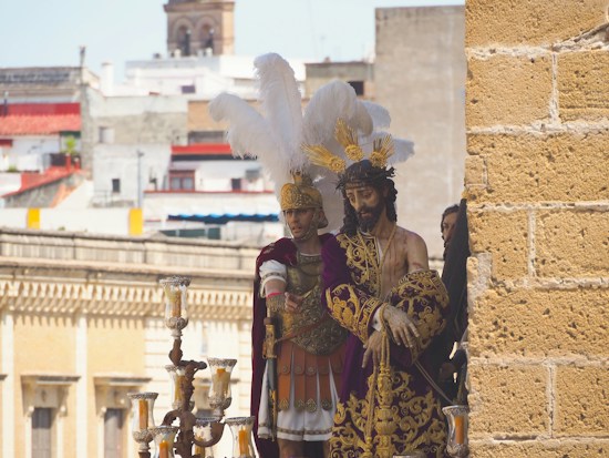 A detailed procession display featuring religious statues dressed in ornate garments. The central figure wears a purple robe with gold embroidery, crowned with a headpiece of feathers, symbolizing a sacred or historical event. A Roman soldier figure is adorned in armor with a striking feathered helmet. The background reveals a cityscape with a mix of modern and traditional architecture.