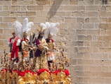 A richly decorated religious procession display featuring several human figures dressed in elaborate, historically-styled garb. The figures are adorned with intricate armor and colorful robes, surrounded by ornate golden decorations and red floral arrangements. White feathers are prominently mounted on some of the headdresses, and an ancient stone wall serves as the backdrop.