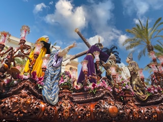 A detailed and ornate religious procession float with human-like figures dressed in vibrant, historical clothing. The float is adorned with intricate carvings, flowers, and lanterns. Palm trees and a bright blue sky with some clouds are visible in the background.