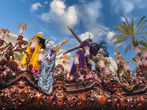 A detailed and ornate religious procession float with human-like figures dressed in vibrant, historical clothing. The float is adorned with intricate carvings, flowers, and lanterns. Palm trees and a bright blue sky with some clouds are visible in the background.