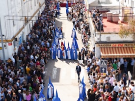 A procession is taking place on a street lined with spectators. Participants in blue and white robes with pointed hoods form a central line. A larger float or object adorned with red cloth is carried further along the route. The spectators are densely packed, creating a festive and solemn atmosphere.