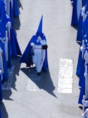 A group of individuals dressed in blue and white robes with pointed hoods walking in a procession. They are carrying staffs and walking on a street with visible pedestrian crossing lines.