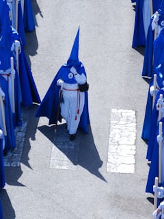 A group of individuals dressed in blue and white robes with pointed hoods walking in a procession. They are carrying staffs and walking on a street with visible pedestrian crossing lines.