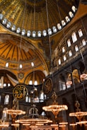 An intricately designed interior featuring a large, ornate dome with geometric and floral patterns. The space is adorned with several large chandeliers emitting a warm glow, and there are circular plaques with Arabic calligraphy hanging from the ceiling. Arched windows allow light to filter in, highlighting the detailed mosaics and structural elements.