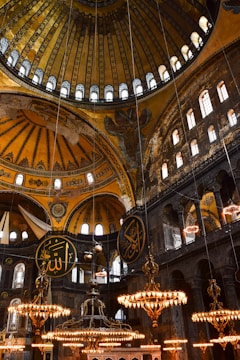 An intricately designed interior featuring a large, ornate dome with geometric and floral patterns. The space is adorned with several large chandeliers emitting a warm glow, and there are circular plaques with Arabic calligraphy hanging from the ceiling. Arched windows allow light to filter in, highlighting the detailed mosaics and structural elements.