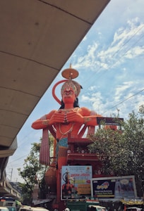 A large statue of a deity with orange skin and a decorated headdress stands prominently in an urban setting. The statue is surrounded by trees and several signs and billboards are visible at its base. A section of an overhead structure and a cloudy sky can be seen in the background.