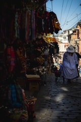 A cobblestone street in a vibrant market area with colorful textiles and masks hanging on display. A person dressed in white with a wide-brimmed hat walks away, pushing a cart. The street is lined with various stalls under bright daylight, and overhead wires crisscross the sky.