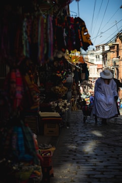 A cobblestone street in a vibrant market area with colorful textiles and masks hanging on display. A person dressed in white with a wide-brimmed hat walks away, pushing a cart. The street is lined with various stalls under bright daylight, and overhead wires crisscross the sky.