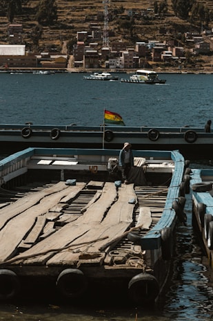 A wooden platform floats on a body of water near the shore, with a small flag fluttering in the breeze. In the background, a town with numerous buildings and houses lines the water's edge, while some boats sail on the water. A person wearing a hat stands on the platform, surrounded by a serene and rustic atmosphere.