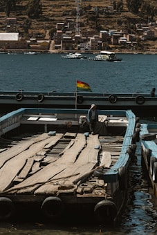 A wooden platform floats on a body of water near the shore, with a small flag fluttering in the breeze. In the background, a town with numerous buildings and houses lines the water's edge, while some boats sail on the water. A person wearing a hat stands on the platform, surrounded by a serene and rustic atmosphere.