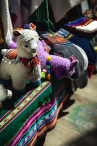 A display featuring colorful, handcrafted llama toys made of fabric with intricate details. The llamas have woolly textures and are adorned with multicolored tassels and traditional patterns. They are set against a vibrant backdrop of textiles, including a deep green cloth with stripes of bright pink, blue, and white.