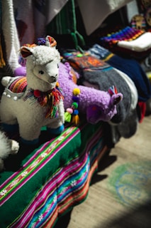 A display featuring colorful, handcrafted llama toys made of fabric with intricate details. The llamas have woolly textures and are adorned with multicolored tassels and traditional patterns. They are set against a vibrant backdrop of textiles, including a deep green cloth with stripes of bright pink, blue, and white.