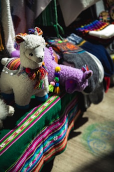 A display featuring colorful, handcrafted llama toys made of fabric with intricate details. The llamas have woolly textures and are adorned with multicolored tassels and traditional patterns. They are set against a vibrant backdrop of textiles, including a deep green cloth with stripes of bright pink, blue, and white.