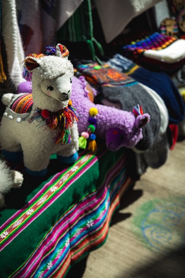 A display featuring colorful, handcrafted llama toys made of fabric with intricate details. The llamas have woolly textures and are adorned with multicolored tassels and traditional patterns. They are set against a vibrant backdrop of textiles, including a deep green cloth with stripes of bright pink, blue, and white.
