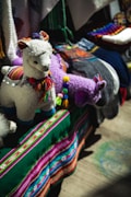 A display featuring colorful, handcrafted llama toys made of fabric with intricate details. The llamas have woolly textures and are adorned with multicolored tassels and traditional patterns. They are set against a vibrant backdrop of textiles, including a deep green cloth with stripes of bright pink, blue, and white.