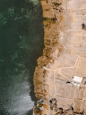 An aerial view of a coastal area featuring a dark green water body on the left side and arid land formations on the right. The shoreline is irregular, with patches of vegetation and exposed earth. There are some rectangular plots of land, suggesting agriculture or construction, along with a few buildings present on the right.