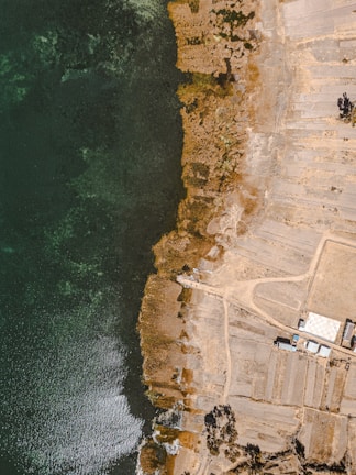 An aerial view of a coastal area featuring a dark green water body on the left side and arid land formations on the right. The shoreline is irregular, with patches of vegetation and exposed earth. There are some rectangular plots of land, suggesting agriculture or construction, along with a few buildings present on the right.
