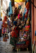 A bustling market scene with a narrow street lined with vibrant textiles and handmade goods. Various colorful fabric items, such as bags and clothes, are displayed on stands and hung around the stall. A person dressed in traditional attire examines the merchandise.