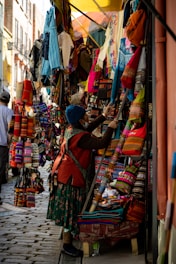 A bustling market scene with a narrow street lined with vibrant textiles and handmade goods. Various colorful fabric items, such as bags and clothes, are displayed on stands and hung around the stall. A person dressed in traditional attire examines the merchandise.